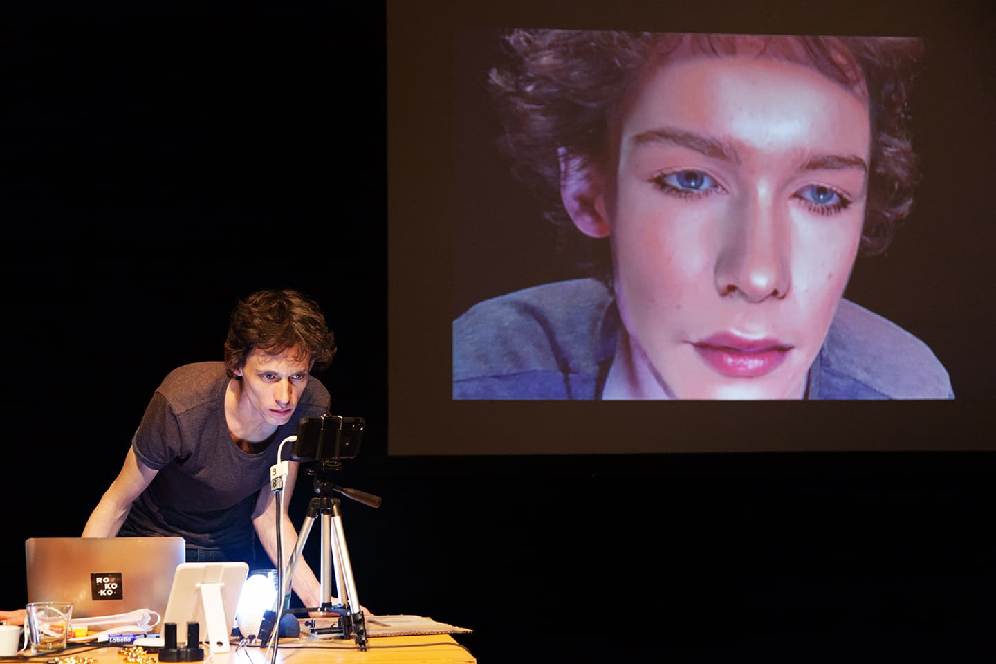 Simon Senn with laptop, microphone and audiovisual equipment at desk. Face of woman, digitally superimposed, on large screen behind him, in the performance Be Arielle F