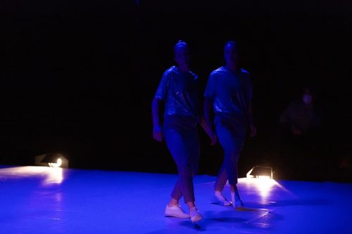 Marion Darova and Martina Apostolova, two women in white sneakers and dark blueish trousers and tops, on a dark bluish stage, with two floor lights behind them, in their stage duet WO MAN