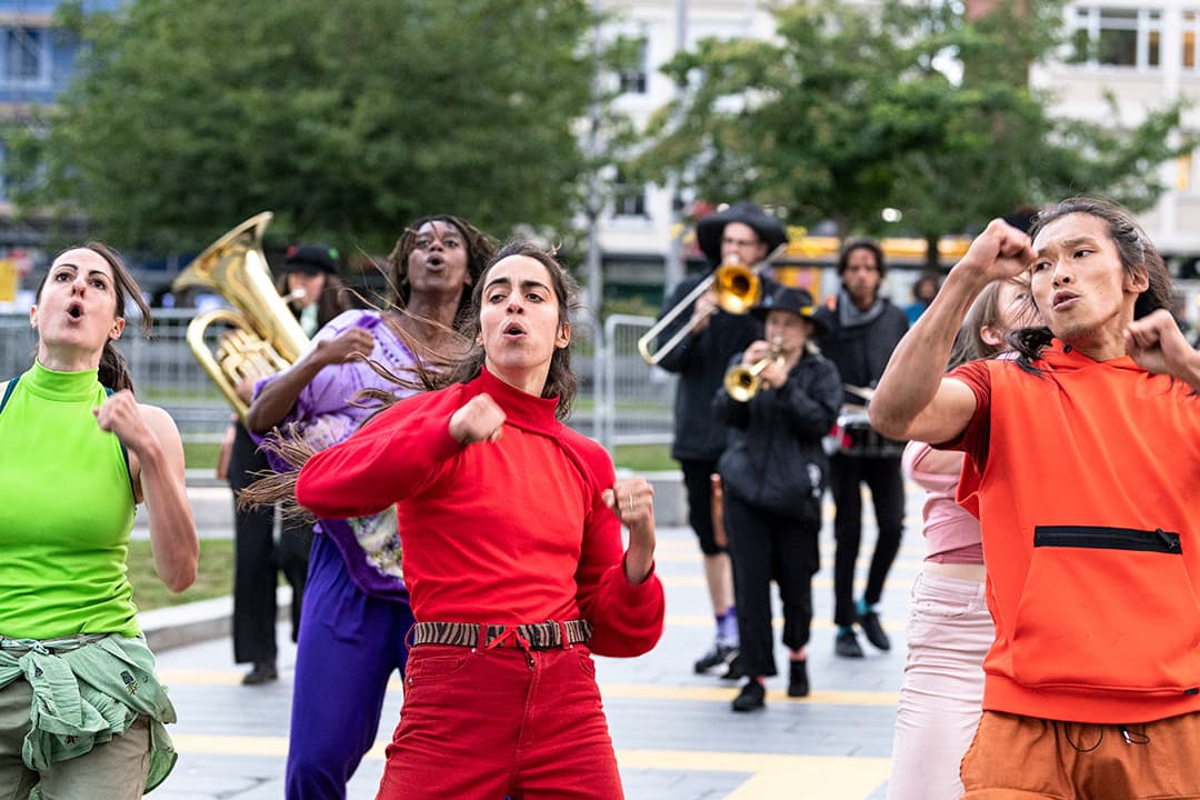 Dancers in bright green, orange, red and purple outfits, in front of a brass band in city square in Woolwich Southeast London. Part of Protein Dance’s guided performance En Route.