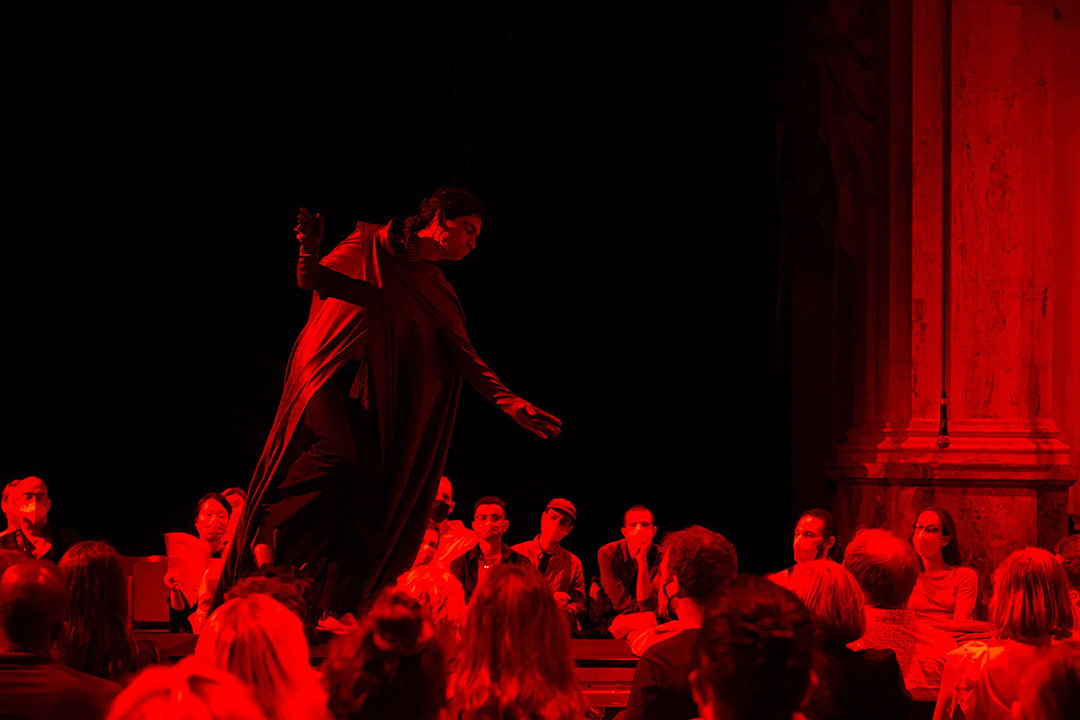 Dark red and black photo of the dance project by PRICE called Melodies are so far my best friend – showing a cloaked man standing on a raised table, surrounded by audience members in Covid facemasks