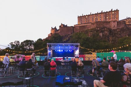 Outdoor audience on socially distanced seats looking at the lit MultiStory stage, Edinburgh Castle in the background