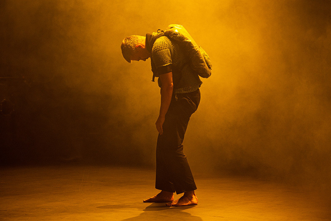 Grief Will Be Our Companion, by Geir Hytten. A barefoot man stands in profile with his head hanging down. On his back is a flat plastic sack. The stage is filled with smoke, the light is orange-yellow