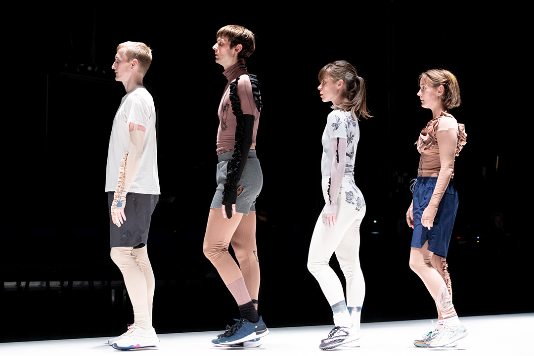 Roza Moshtaghi’s Limbo. A line of four performers, standing in profile. On the left, a man in white t shirt and grey shorts. Next another figure in grey shorts, tan tights and top. Then a oman in white leggings and top with floral black embroidery. Finally another woman in tan tights and top, and blue shorts. All wear trainers.