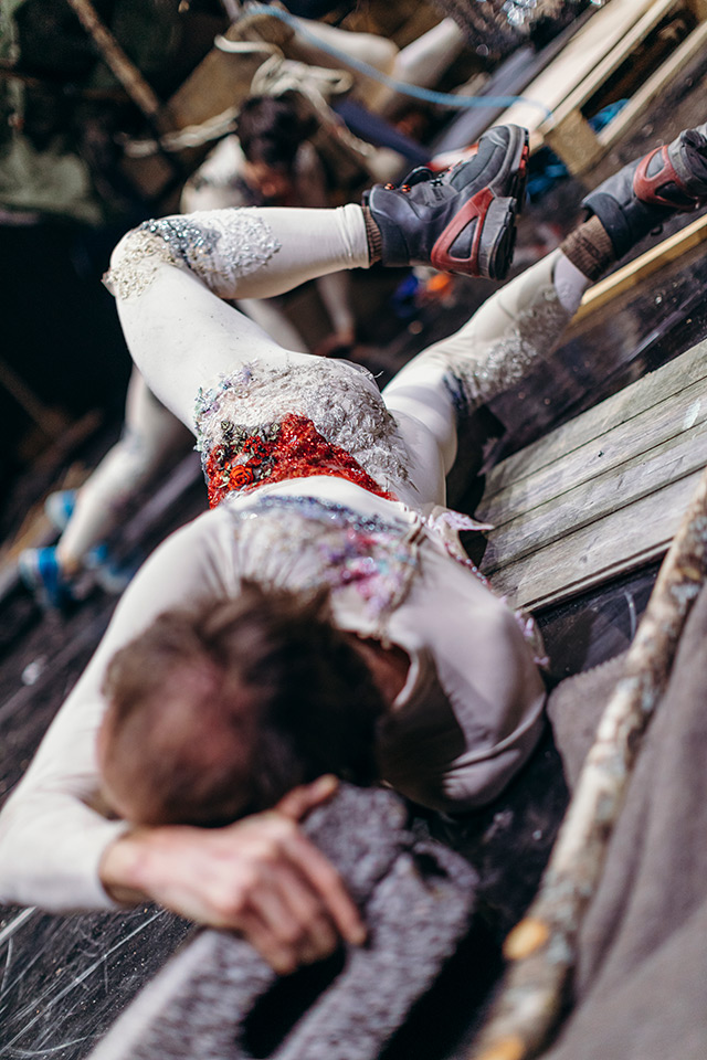 Mia Habib’s How to Die, Inopiné. Man lies face down at an angle across wooden pallets. Wearing a grey unitard embroidered with lichen-like grey and red stitching