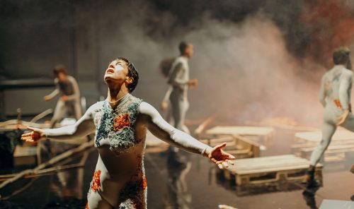 Mia Habib’s How to Die, Inopiné. Woman with cropped hair and outstretched arms is looking up in the foreground. She wears a grey unitard embroidered with lichen-like greys and oranges. In the background are four others, in similar costumes. Wooden pallets lie on the floor and there is smoke in the background.