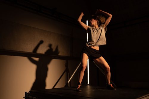 Mette Ingvartsen in The Dancing Public. Woman in trunks and sleeveless top, head thrown back and arms up, on a platform. Behind and to the right of her is her own shadow on the wall