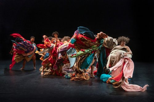 Encantado by Lia Rodrigues. A ragged line of dancers swathed with exuberant multicoloured cloths and fabrics swirling all around them