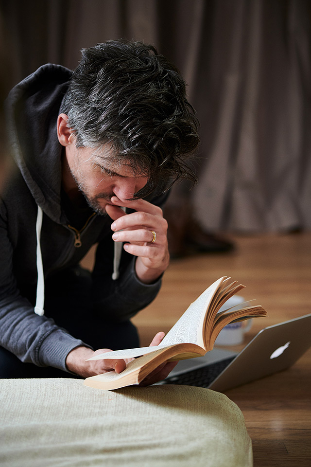 Ben Duke in grey tracksuit top, seated, one hand against his lips as he looks down a book (A Tale of Two Cities) held in the other