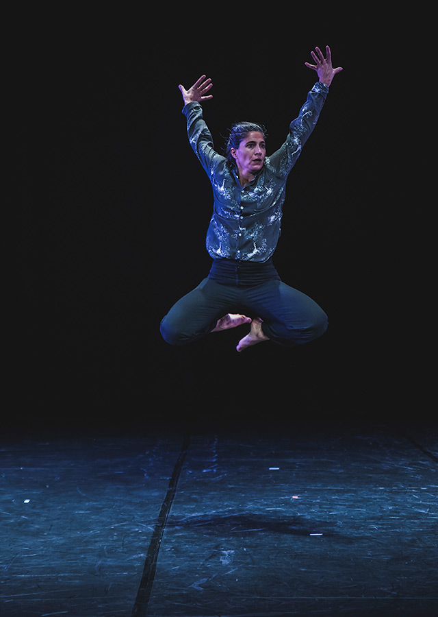 Woman in dark blue shirt and trousers against a dark stage. She is jumping mid-air, with both arms up with fingers spread, and both feet tucked under her