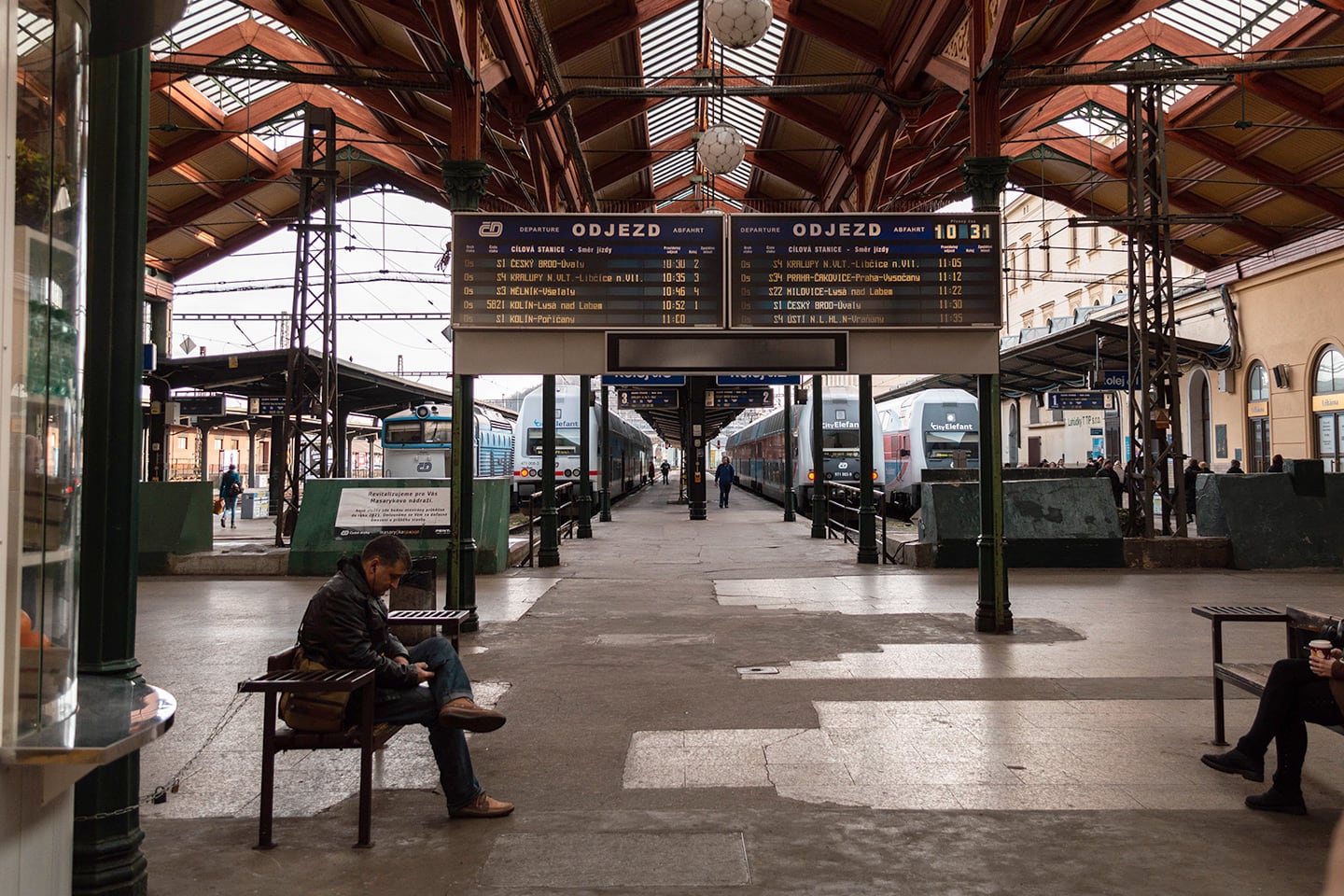 Foreground: man sits on wooden bench in Prague Station. Behind a flipboard showing train times. Behind that, the trains at the platforms.