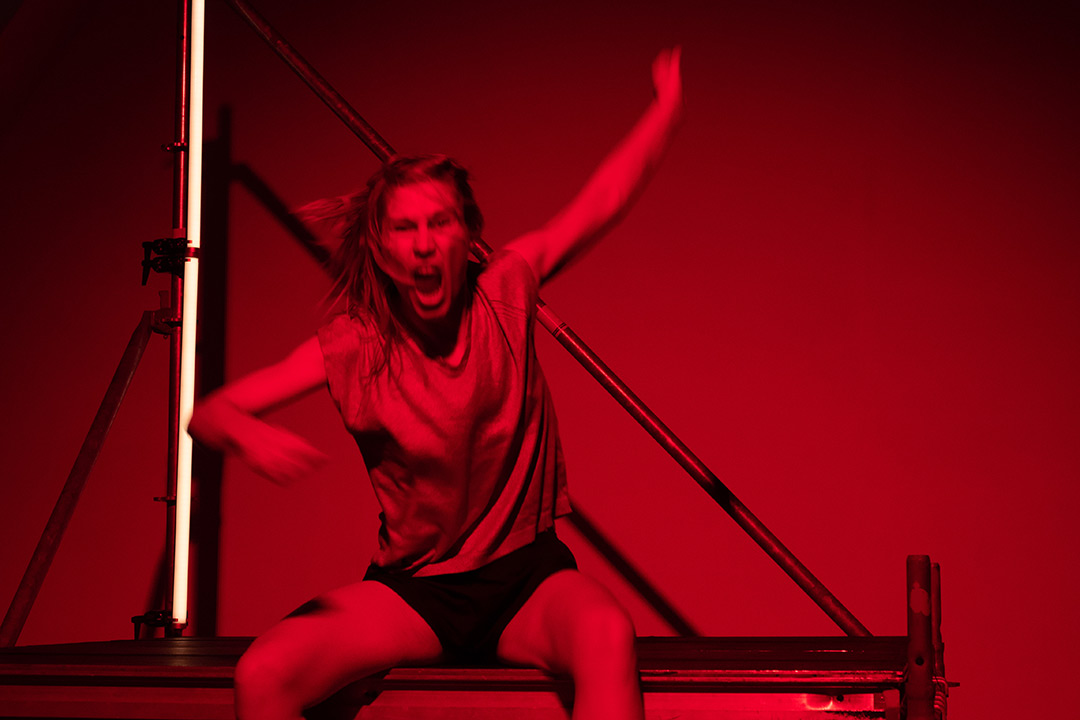 Seated woman in vest and trunks, left arm stretched up, right arm bent forward, mouth open and hair flying. Behind her the diagonal rungs of a lighting rig and a vertical strip light. The whole photo has a deep red colour cast