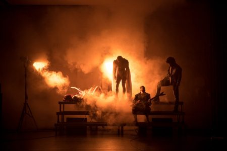 A bench surrounded by dramatic smoke and slanting orange lighting. Dark figures: one guitarist lying down, another sitting, two performers stand on the bench behind them.