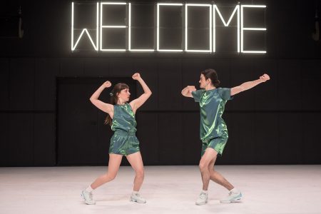 Man and woman in shiny green shirts and shorts face each other in front of a big neon sign which says WELCOME. Compagnie La PP (Romane Peytavin and Pierre Piton) in Farewell Body.