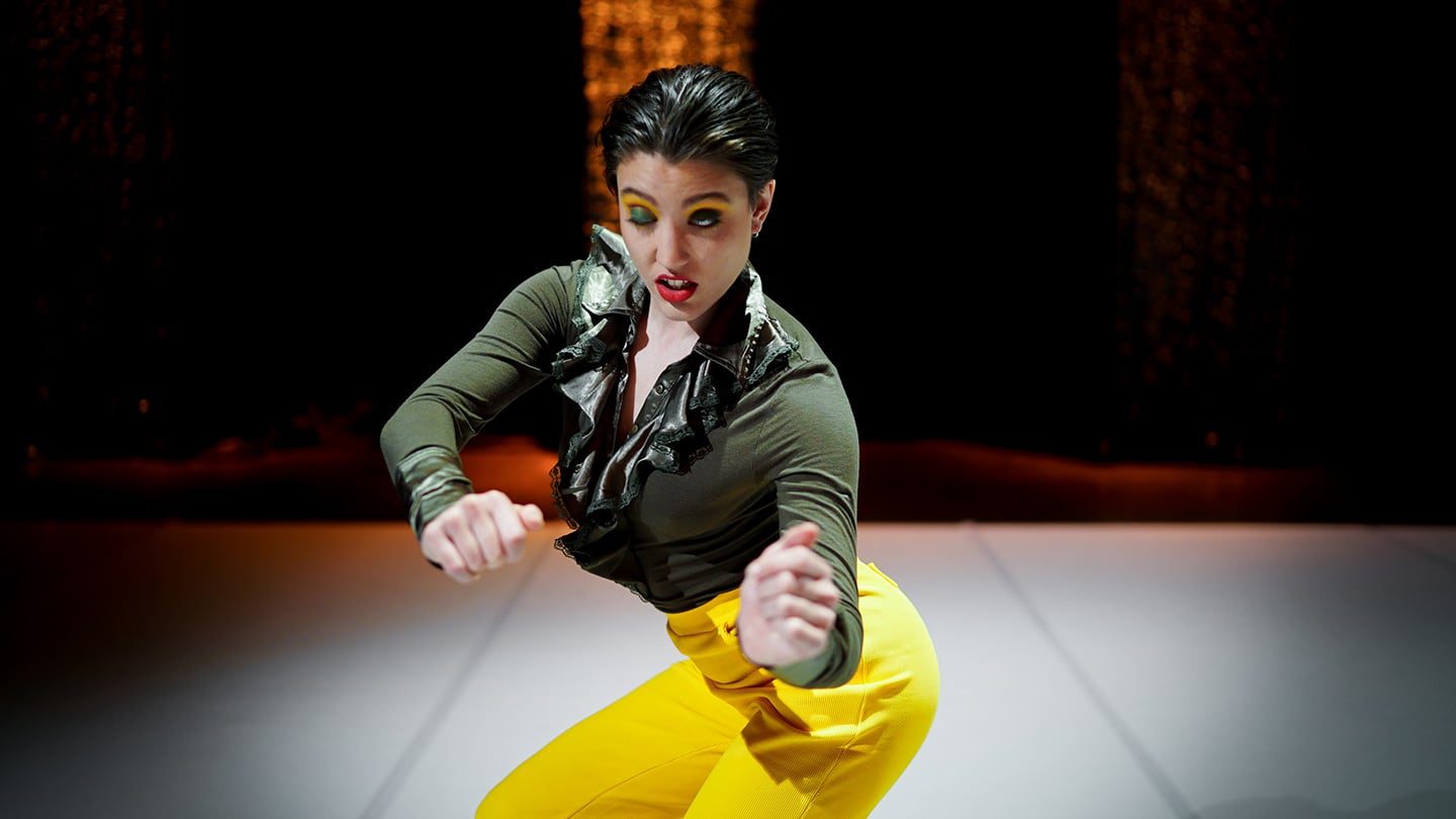 woman in tight olive-green shirt and bright yellow trousers, dark hair slicked and heavy eye makeup, one eye open, one eye closed. She twists towards the viewer, with hands held in fists