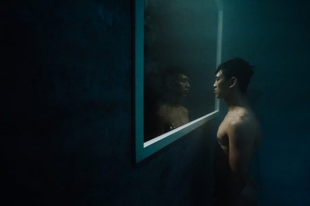 In the darkness, one man looks through a window-like frame at another, as if in a mirror. Both are unclothed.