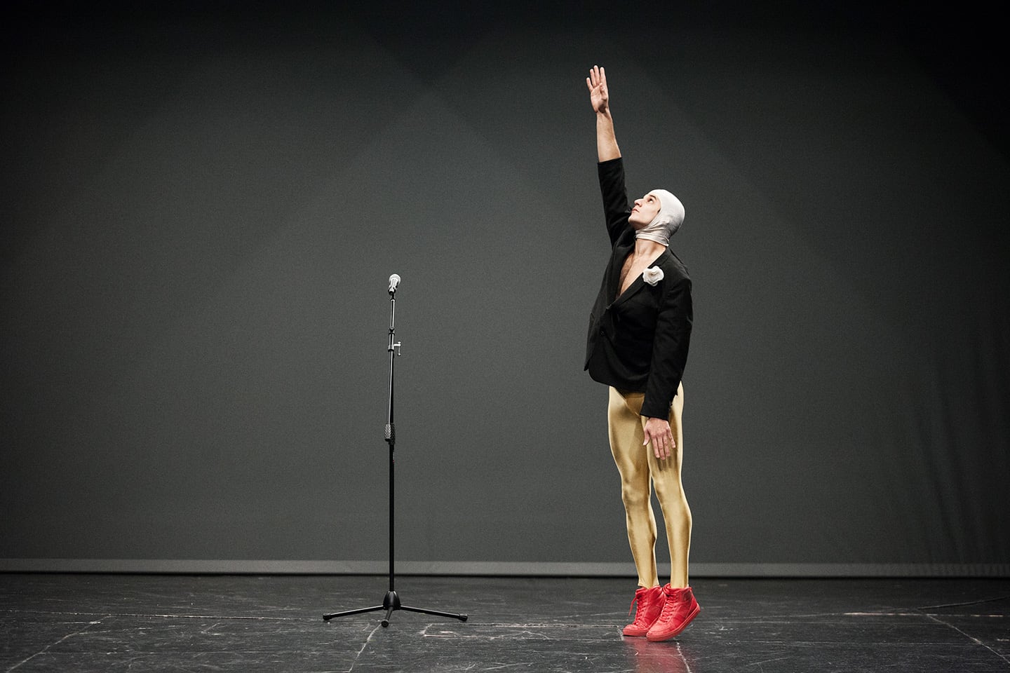 Man in silver balaclava, black jacket, gold tights and red boots, reaches upwards on tiptoe. Next to him is a microphone stand