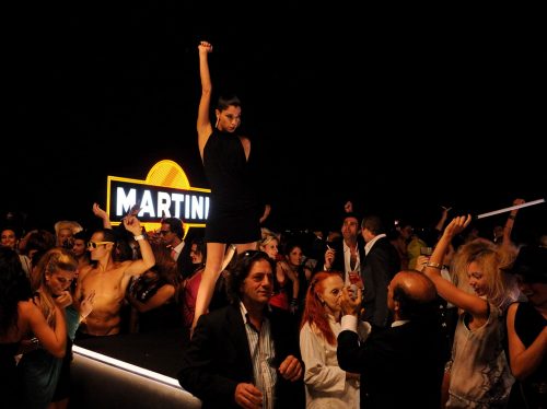 Party scene from Sorrentino’s La Grande Bellezza, young woman stands on a bar with one arm raised, fist clenched. Around her partygoers are revelling, behind her a Martini sign.