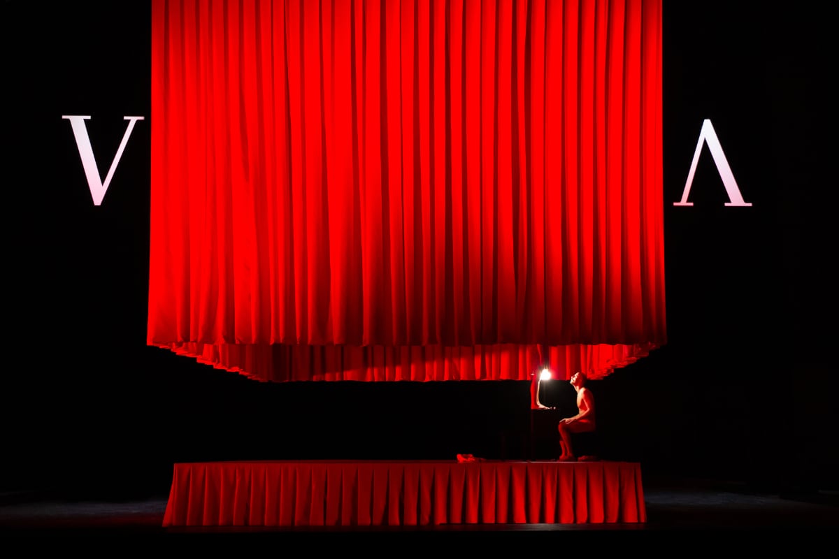 A long red pleated curtain hangs in a square around raised square stage, itself fringed by a pleated red curtain. In the gap between lower and upper curtains, we see setated man looking up, and the feet of a woman dangling down.