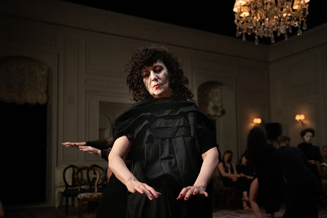 Woman in dark dress, white facepaint and red lips looking at her own hands, held before her. Behind her another per of hands can be seen. In the background are audience members seated in the room, which is a kind of formal drawing room, with chandelier