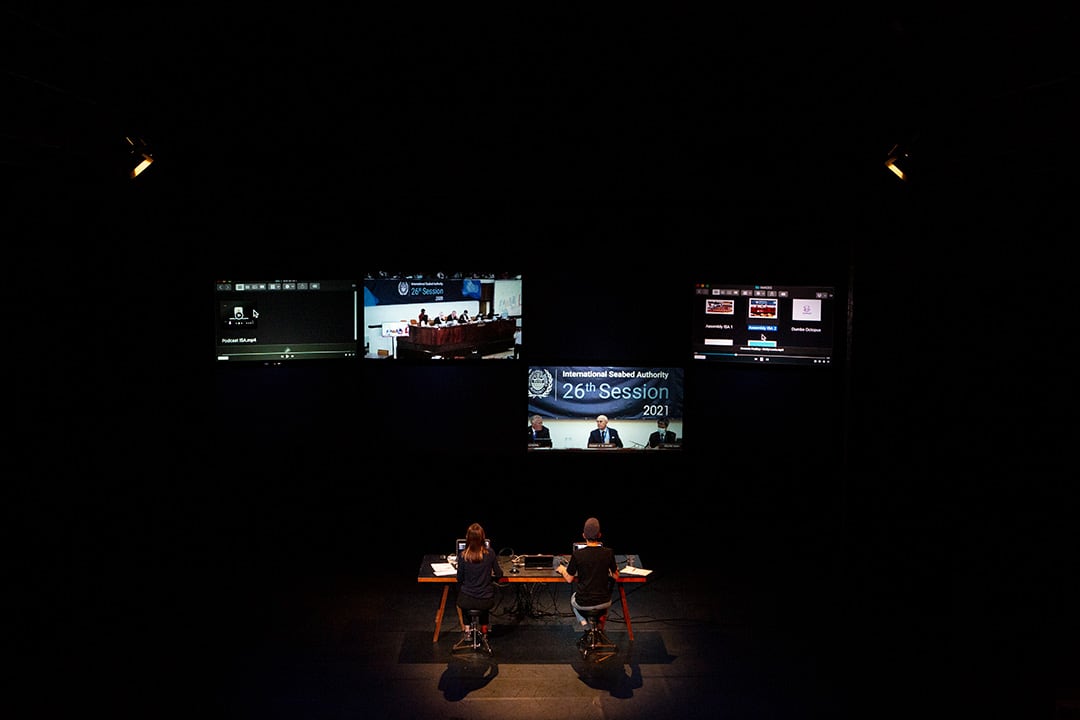 Against a very dark space, four screens are suspended showing computer relays of climate panels, computer filins and folders. Below and in front, two performers sit at a mixing desk with their backs to us