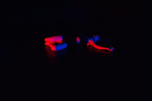 Woman lying twisted on dark floor with dark background, almost as if suspended in air. A red/blue light highlights parts of her legs and one arm.
