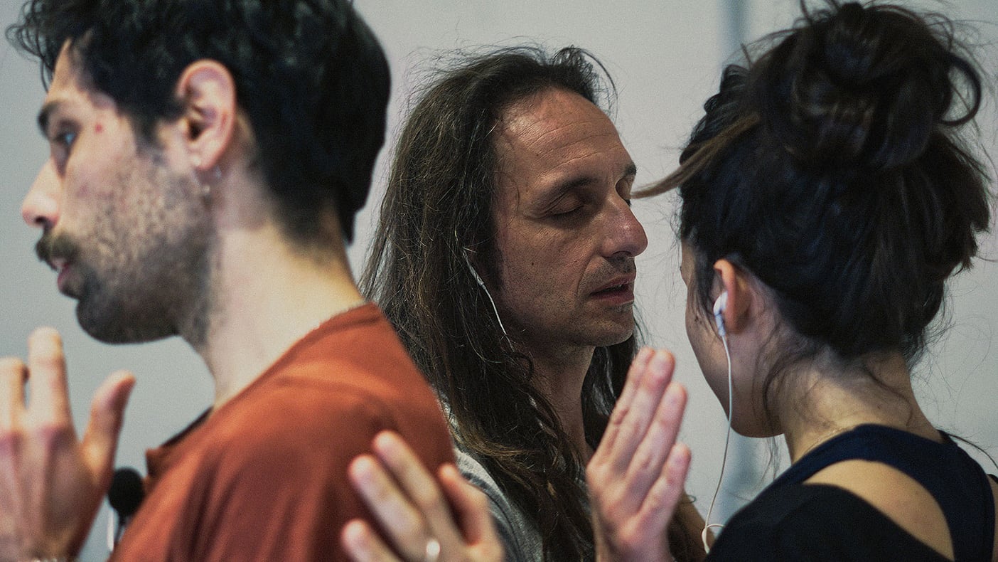 The heads of three people: a young man with stubbly beard and tan top on the left. An older man with eyes closed and long hair in the middle. A woman with tied black hair and black top on the right. All wear earphones, and all seem lost in the world they are listening to, their focus turned within though their hands reach towards each other