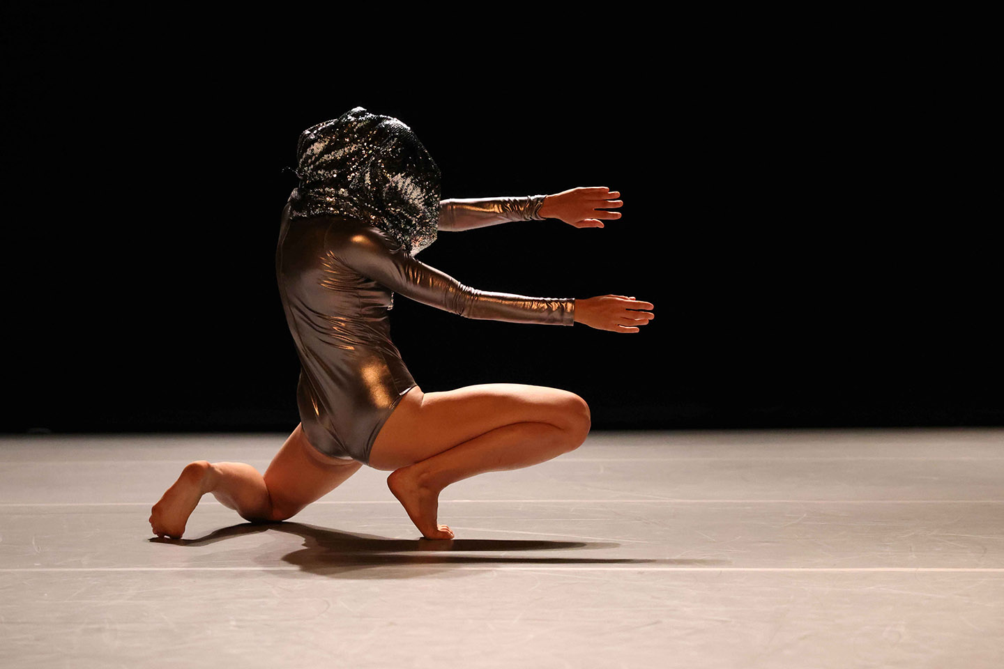 Frieze-like picture of woman in shiny silver leotard crouched on the floor, arms extended in front of her. Covering her whole head is a large bulbous silver mask. The look is mythical/futuristic