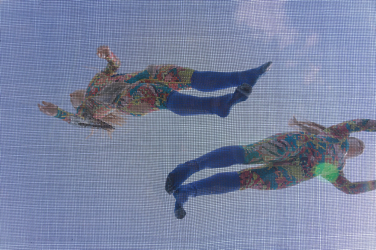 Looking upwards through a trampoline mesh, two colourfully dressed figures are outlined like strange birds against a blue sky