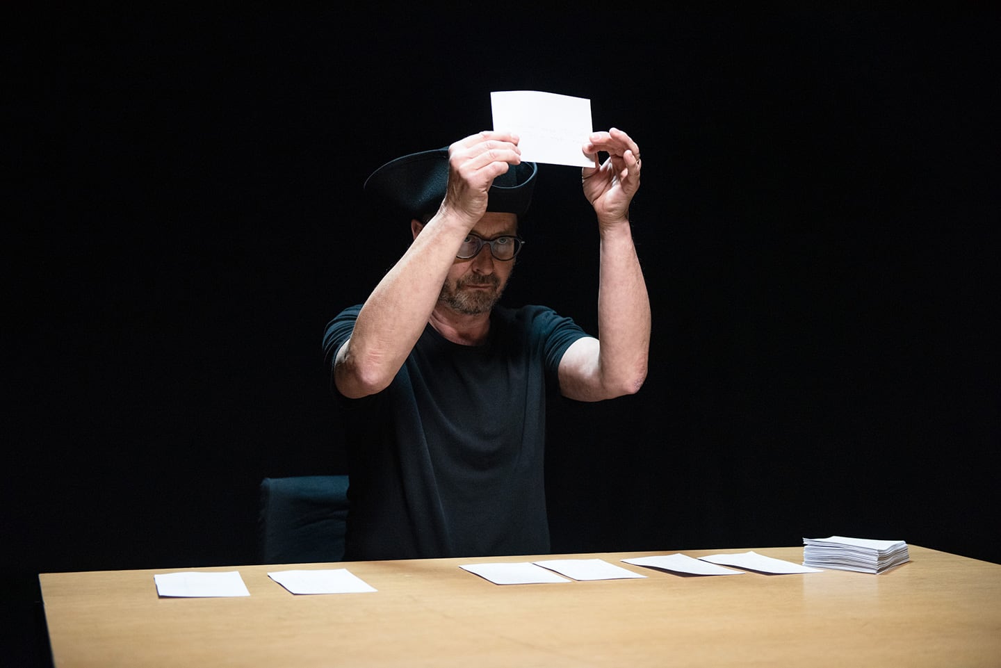Bespactacled man in pirate hat holds up a blank card from a row of blank cards dealt on the table before him