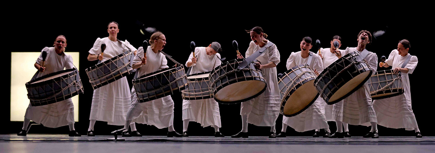 A ragged row of nine women in long white dresses, each banging a huge horizontal drum