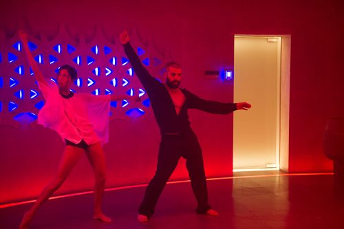 Woman in gauzy top and underwear, man in dark trousers and open shirt, each in disco-pose with one arm up and the other out. Behind them are neon blue lights, and a yellow door, and the whole scene is soaked in dark red light.