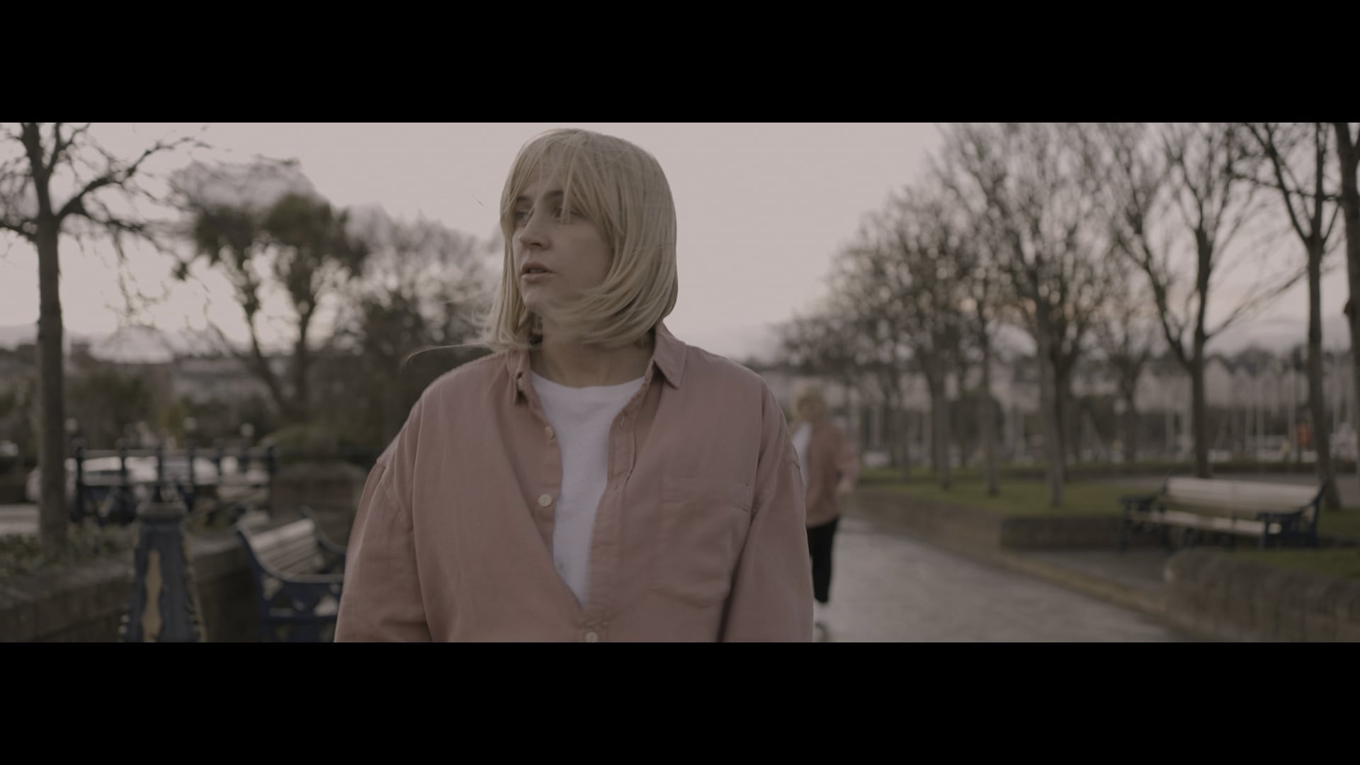 Woman in salmon shirt and blonde wig in the foreground, behind her, blurred is another in the same outfit. Both pictured against a tree-lined park road on a grey day