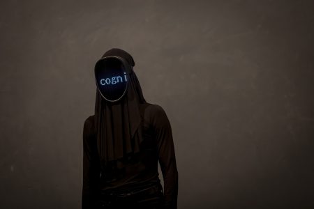 A female figure completely cased in black clothes with a mask over her face on which a digital word is beamed (the letters "cogni" suggesting the beginning of the word "cognition")