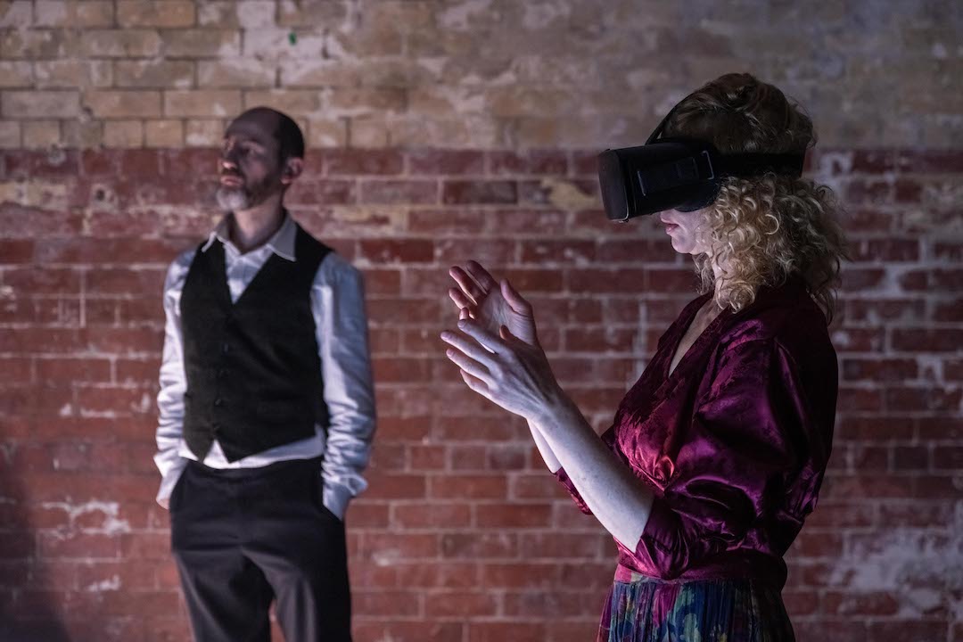 Woman wearing VR headset looks to the right, with hands raised before her. A man in a waistcoat in the background looks outwards
