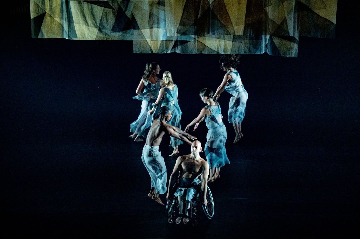 Dancers in loose V-formation with wheelchair user at the front, the others lined behind with arms loosely stretched forward. Above them hanging transparent cloths with geometric patterns echo their loose translucent costumes