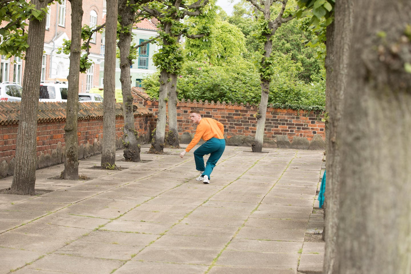 Man in orange top and turquoise trousers is running away down a paved treet lined with green trees.