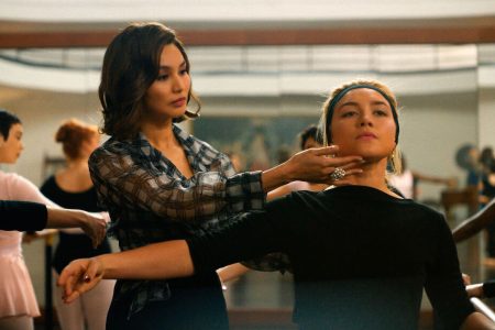 A ballet teacher (Gemma Chan) in checked blouse and ostenatious ring on her finger gently lifts the head of another woman (Florence Pugh) in black, at a ballet class. Behind them, other women (blurred focus) are also at the ballet barres.