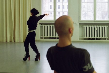 An airy studio space. In the background, a black-clad woman in very high heeled boots faces away, one arms extended towards the windows behind. In the foreground, a bald-headed man, torso only visible in the photo, leans backwards