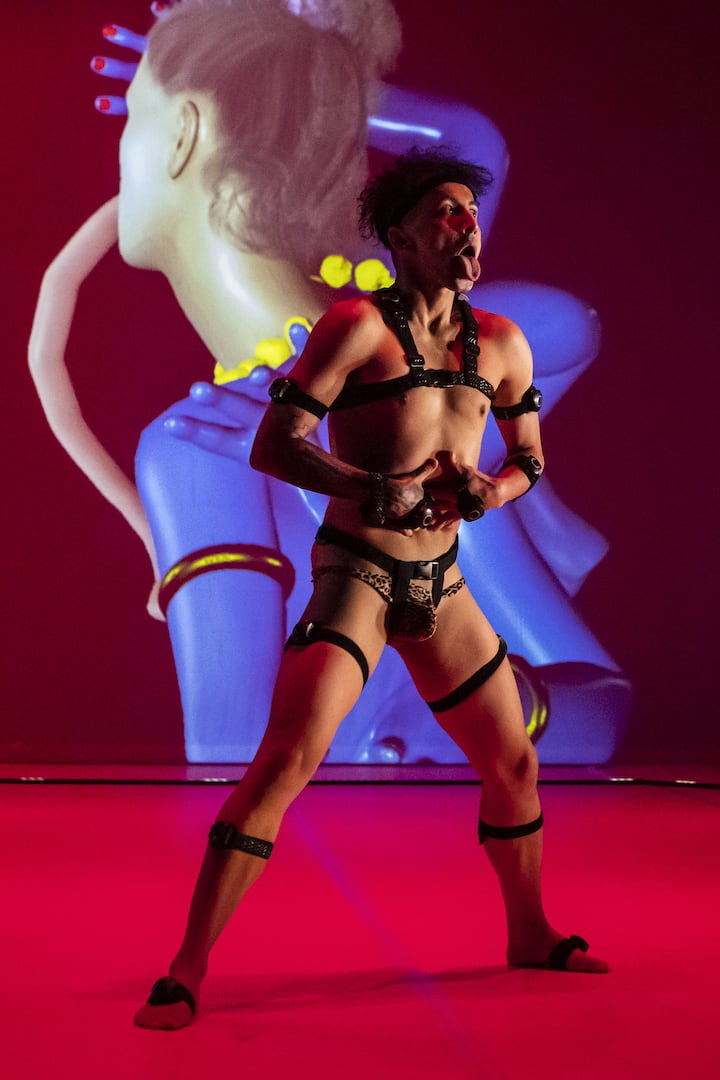 Man in BDSM leather braces on red floor in front of an image of another figure, turned away, in futuristic blue, gold and yellow.