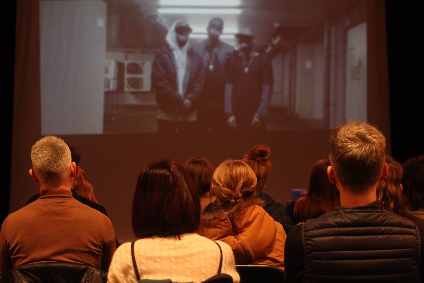 On a blurred background, three black men appear on a screen. In the foreground, we see the backs of the heads of the audience (all white, as far as we can tell) who are watching the screen