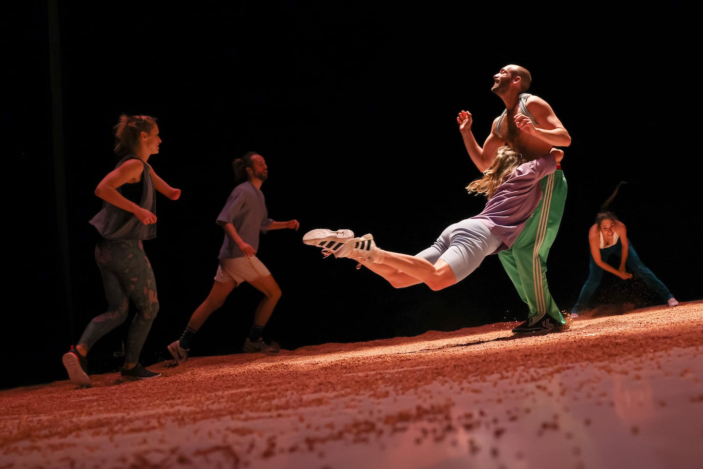 On a tilted floor strewn with reddish grains a woman launches herself headlong and is caught by a man. In the background, other figures are running