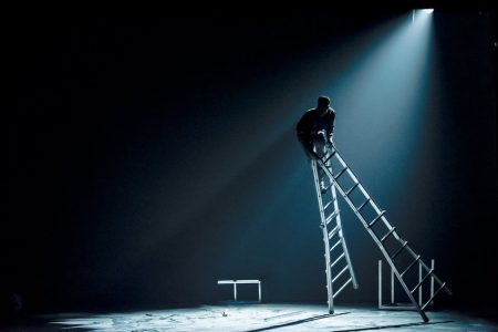 In a slanting spotlight on a shadowed stage, a man is perched perilously on top of a double-sided ladder that is falling
