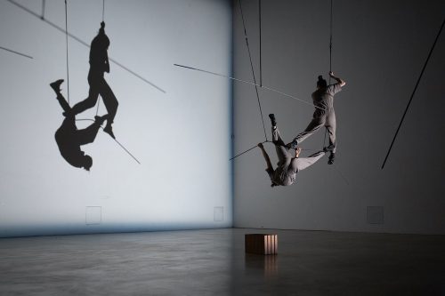 Two figures suspended in the air, one upright, one upside down, balancing on long rods also suspended from the ceiling, in a geometrical but asymmetrical formation. The tone is grey, and behing they their grey shadows are cast against the wall