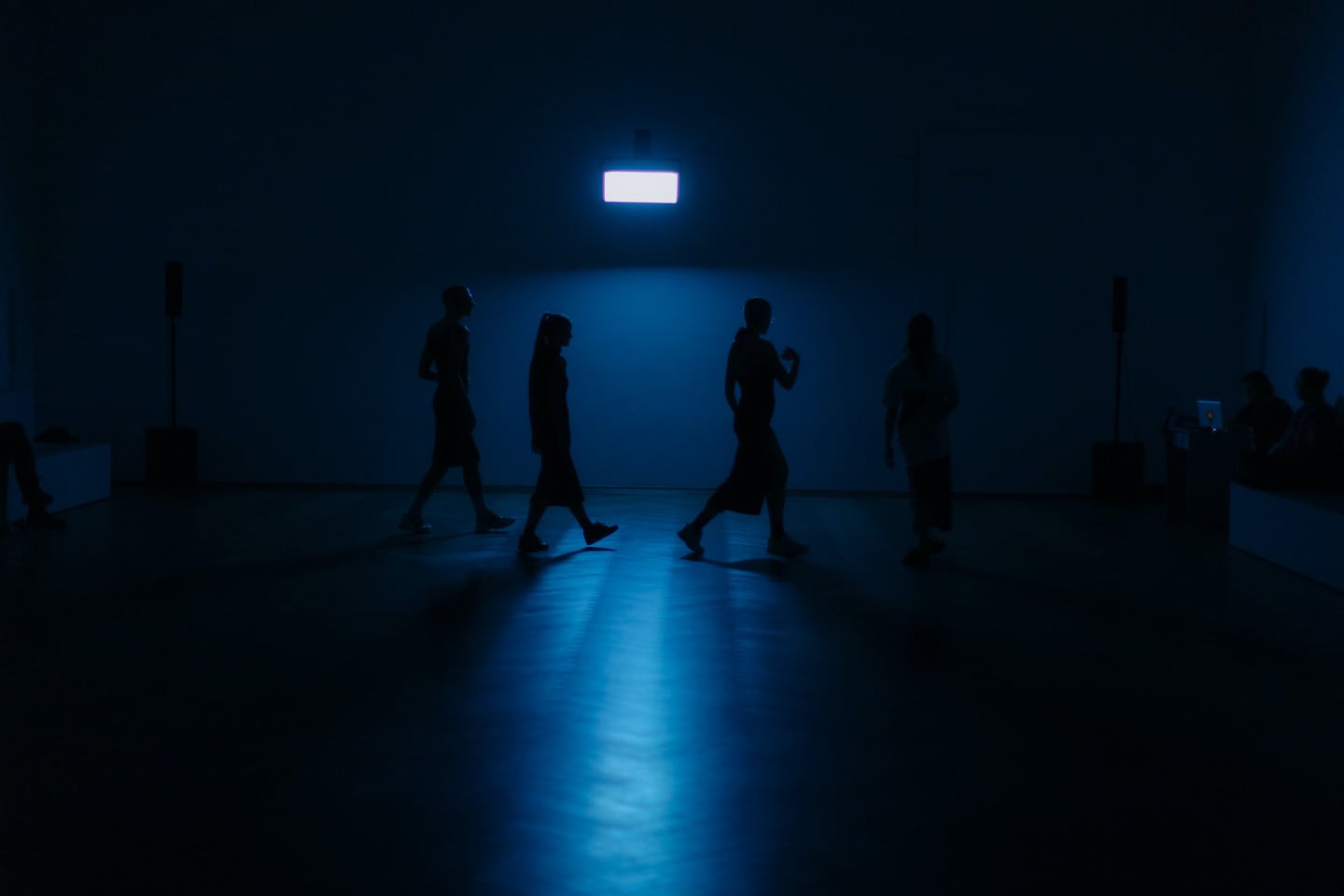 A cavernous dark blue room with a window of white light shows shadowed, silhouetted figures walking in a row across the back