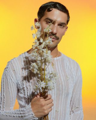 On a bright yellow background, a figure in loos white shirt with dark hair and mustache holds a bunch of white flowers