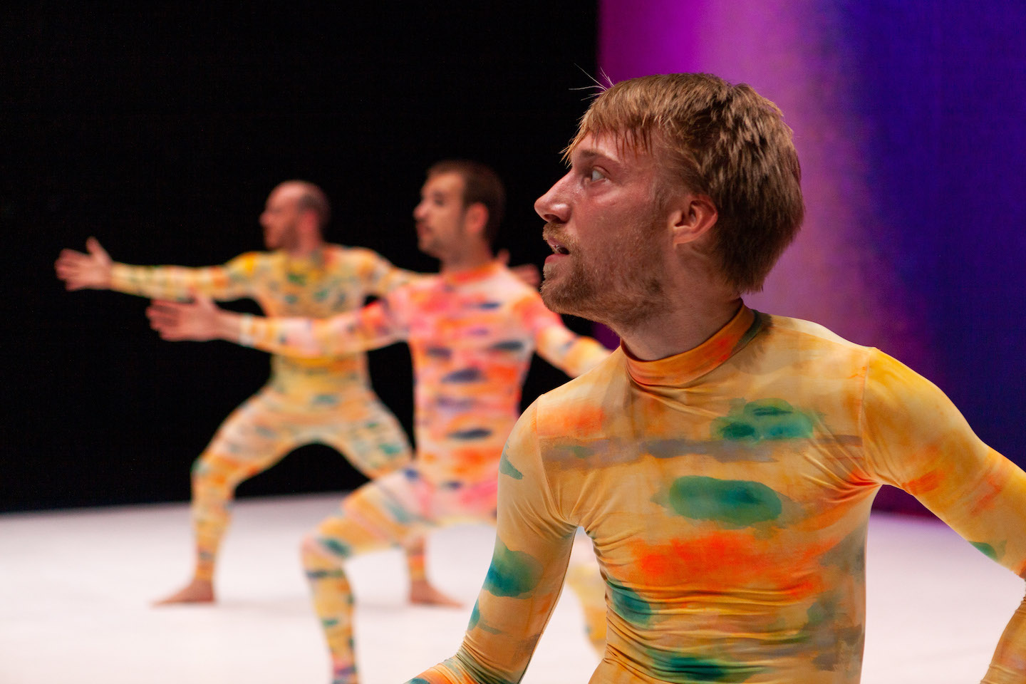 Three men in bright yellow unitards dappled with green, orange and blue, all looking to the left and bent into a wide demi-plié, against a dark purple background. The man in front is in sharp focus with a somewhat alarmed expression, the two behind are blurred.