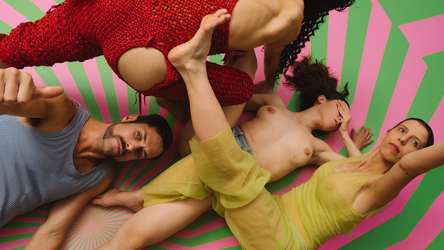 Two men and two women form a crisscross of coloured bodies (red, blue, greem) on pink and green op-art floor.