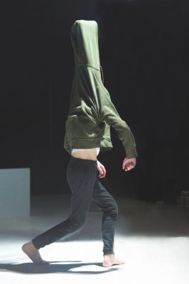 A man walking in a green hoodie, with the hood stretched high up over a hidden block balanced on his head. From Nice Trip, by Mathieu Desseigne-Ravel and Michel Schweizer.