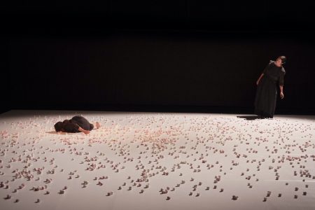 On a white stage strewn with marshmallows, a small figure (Chiara Bersani) lies down in one corner, while from another a standing figure (Elena Sgarbossa) moves towards her.