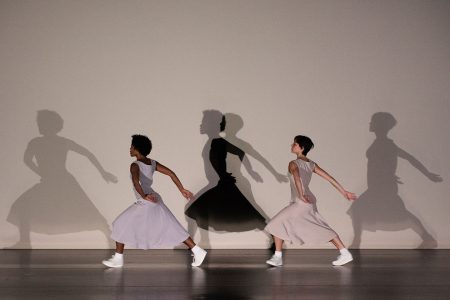 Two women in flowing dresses and white sneakers, stepping sideways in parallel positions. Behind them, their shadows multiply and overlap their outlines